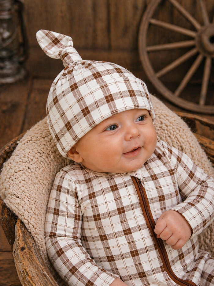 Smiling baby in a plaid outfit and hat, sitting in a cozy basket with rustic decor in the background.