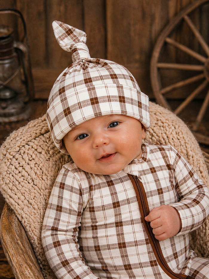 Smiling baby in a plaid outfit and hat, sitting in a cozy setting with wooden decor.
