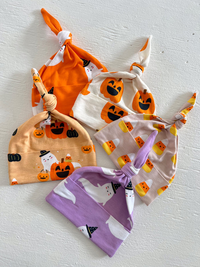 Four playful Halloween-themed baby hats in orange, white, and purple with ghost and pumpkin designs.