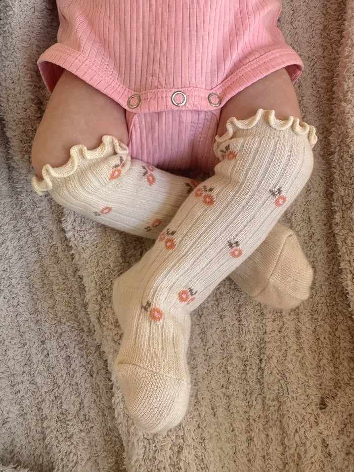 Infant's legs in floral knee-high socks, wearing a pink onesie, resting on a cozy textured blanket.