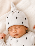 Sleeping baby wearing a light gray hat with black lightning bolt patterns, resting on a soft blanket.