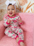 Smiling baby in floral pajamas sitting on a pink chair with a large white bow headband.