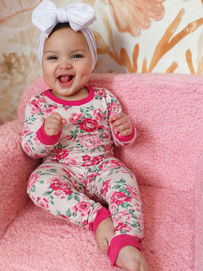 Smiling baby in floral pajamas sitting on a pink chair with a large white bow headband.