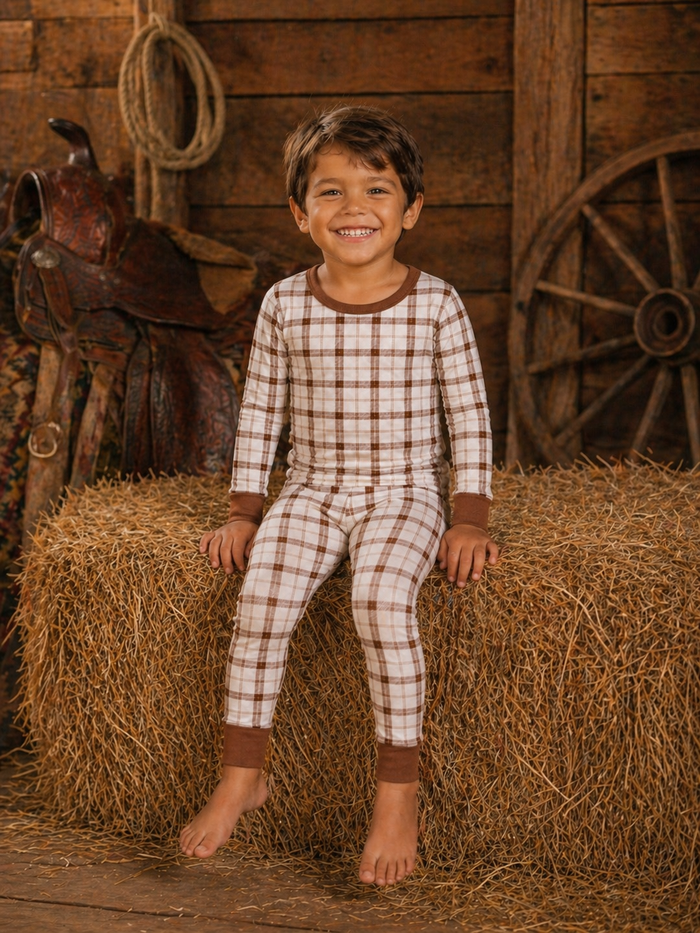 Smiling boy in plaid pajamas sitting on a hay bale, with a rustic barn backdrop and saddle nearby.