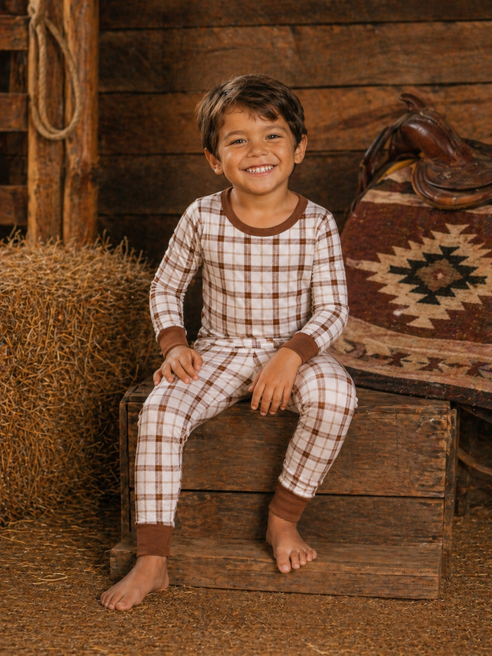 Smiling boy in plaid pajamas sitting on a wooden crate with hay and rustic decor in the background.