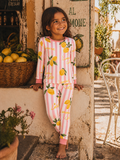 Smiling girl in lemon-patterned pajamas stands near a stone wall and lemon basket, enjoying a sunny day outdoors.