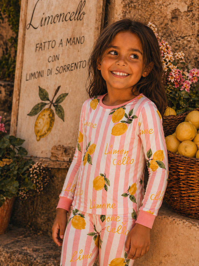 Smiling child in lemon-themed pajamas near a lemon sign and basket of lemons, surrounded by flowers.