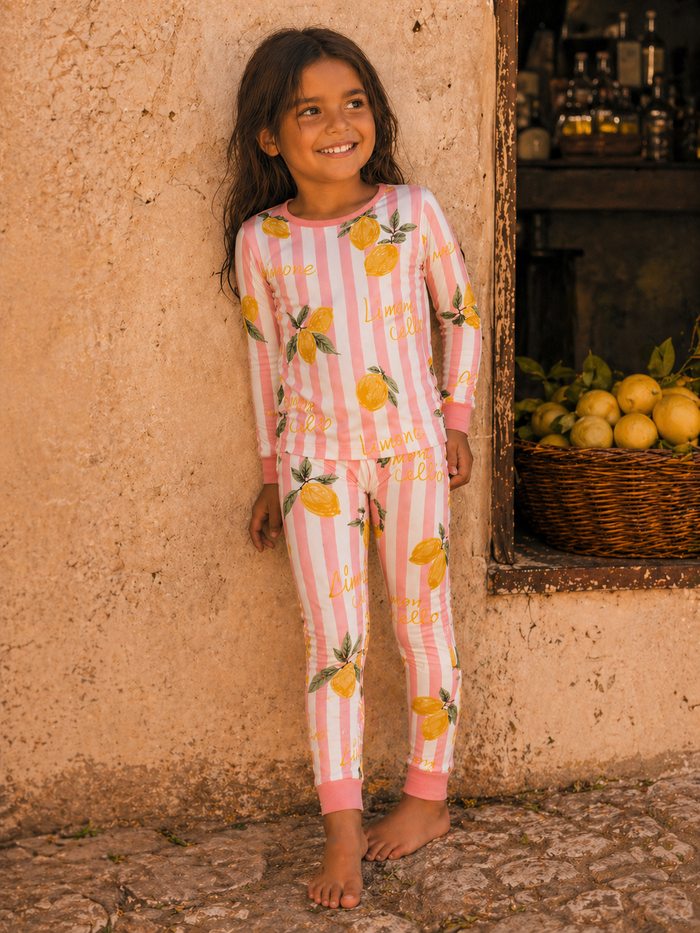 Smiling girl in lemon-patterned pajamas stands against a textured wall, barefoot, with a basket of lemons nearby.