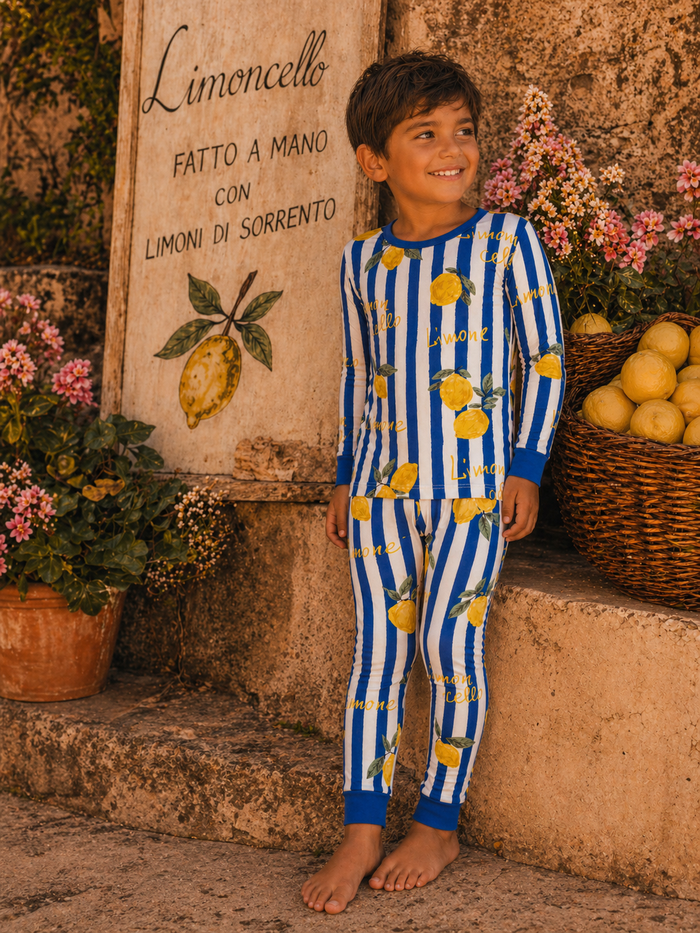 Child in lemon-print pajamas smiles by a Limoncello sign with flowers and a basket of lemons in the background.