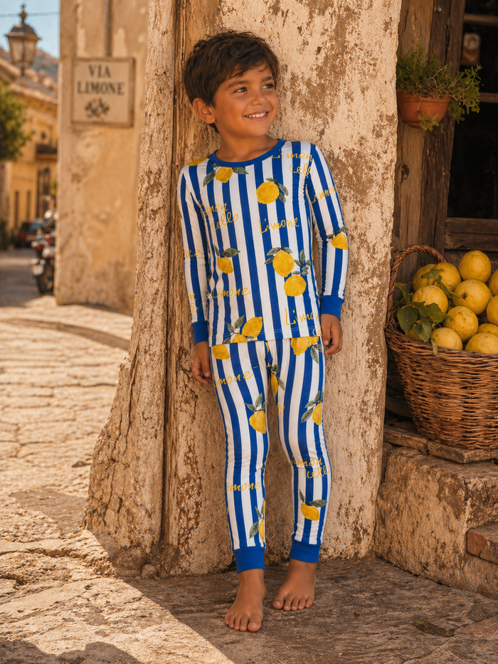 Boy in lemon-patterned pajamas smiles, leaning against a rustic wall with a basket of lemons nearby.