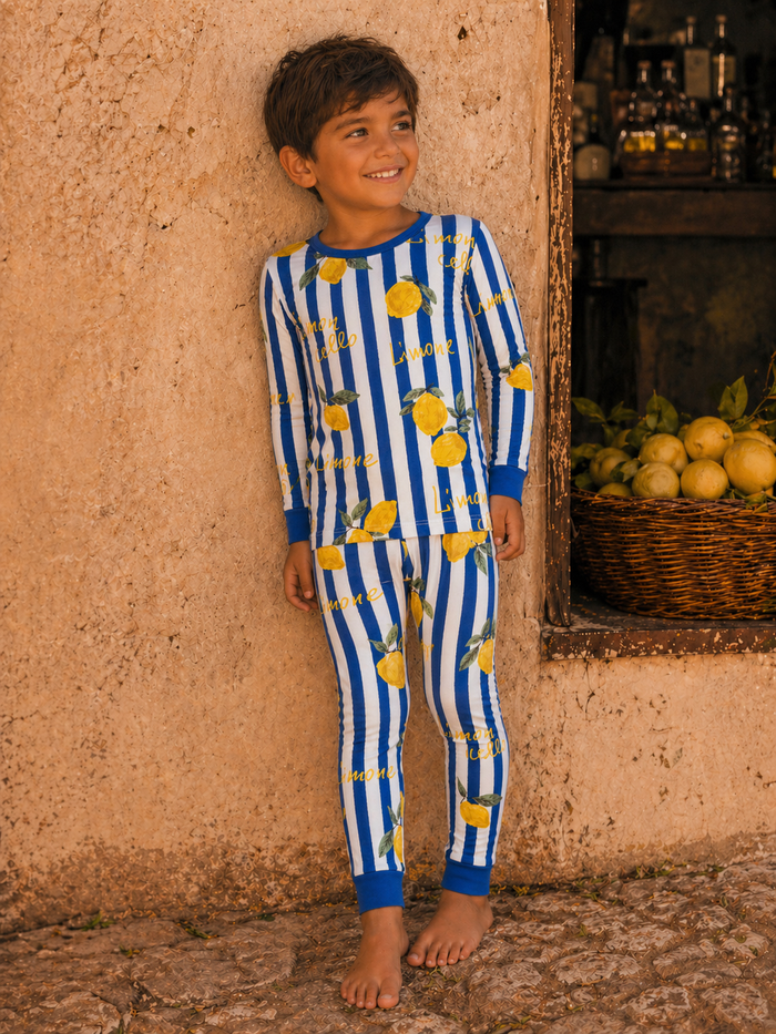 Smiling boy in lemon-patterned pajamas stands barefoot against a textured wall, with a basket of lemons nearby.