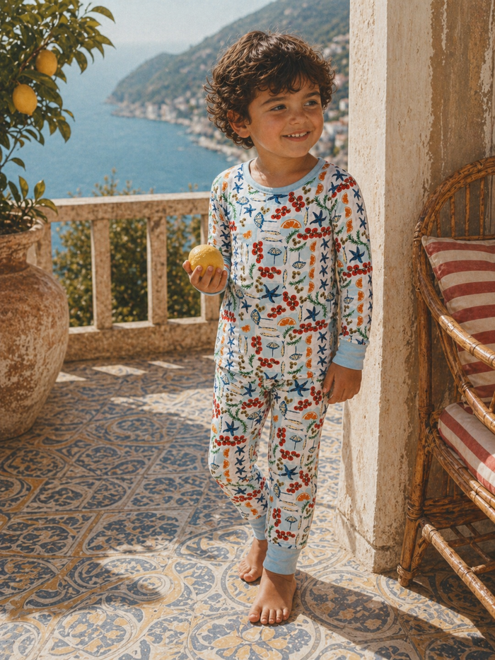 Child in colorful pajamas holds a lemon, smiling on a sunlit terrace overlooking the sea.