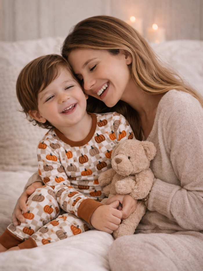 Smiling woman and child in cozy pajamas, holding a teddy bear, seated together on a bed.