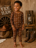 Smiling child in brown pajamas with stars and moons, standing in a rustic setting with cowboy boots and a lantern.