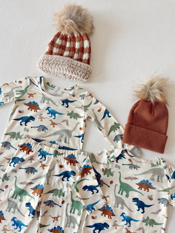 Dinosaur-patterned pajamas with two matching knitted hats, one with a pom-pom, on a white background.