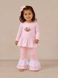 Smiling girl in pink striped outfit with a football graphic and ruffled pants, standing against a neutral background.