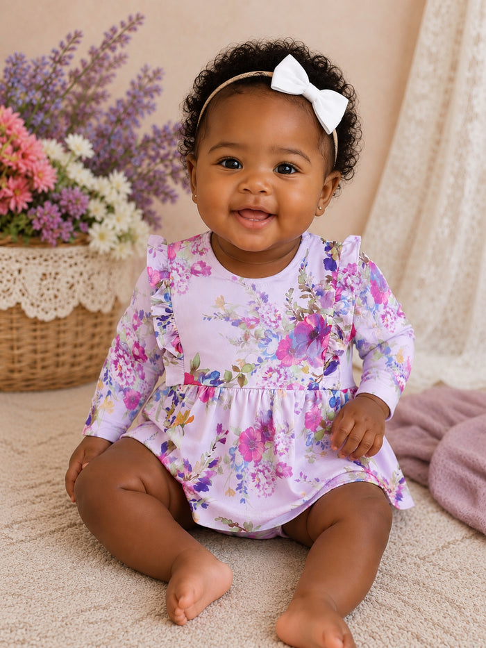 Smiling baby girl in a floral dress and white bow, sitting on a soft surface with flowers in a basket nearby.