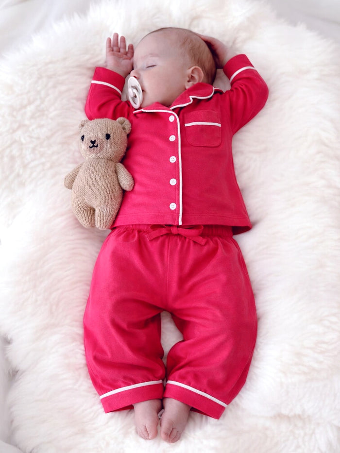 Baby in red pajamas sleeping on a fluffy white blanket, holding a teddy bear.