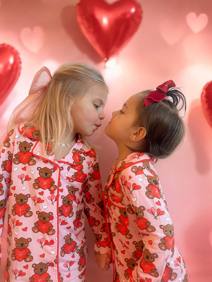 Two young girls in matching bear print pajamas kiss, surrounded by heart-shaped balloons on a pink backdrop.