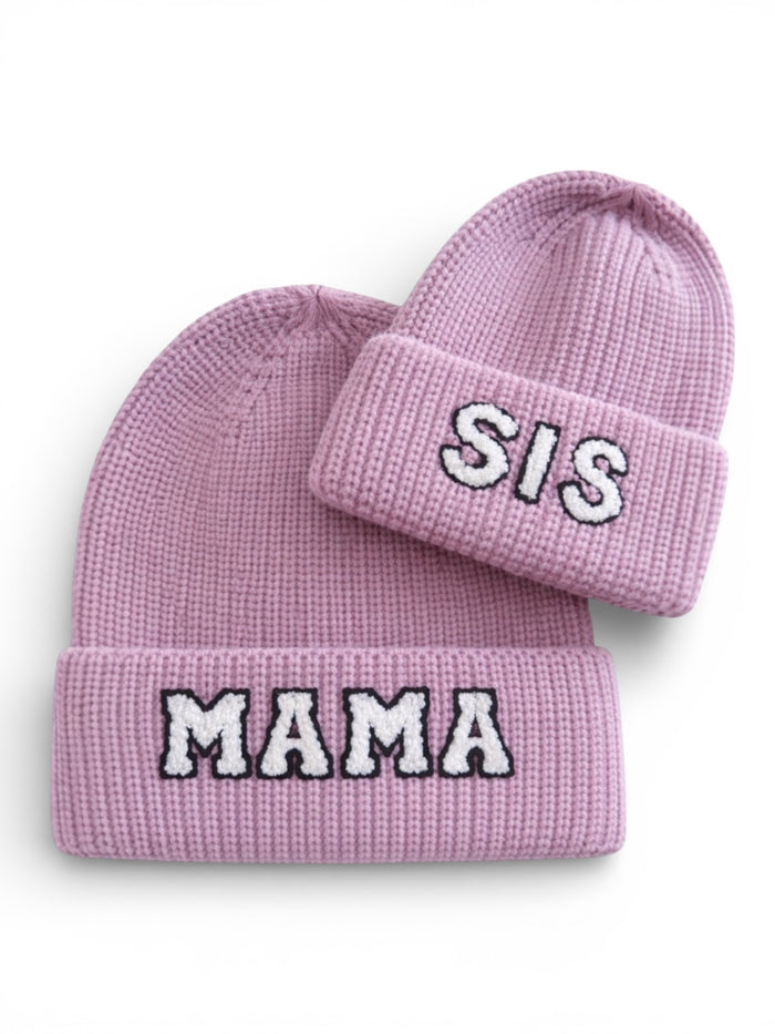 Two pink knitted beanies featuring the words "MAMA" and "SIS" in bold, contrasting lettering.