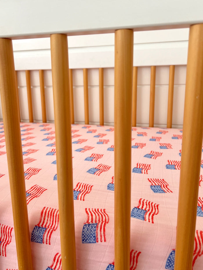 Crib with wooden slats and pink fabric featuring a repeated American flag pattern.