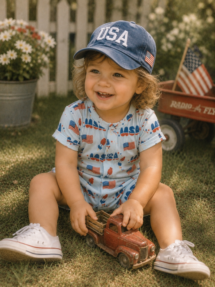 Smiling child in a USA cap playing with a toy truck on grass, surrounded by flowers and a wagon.