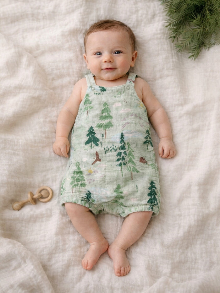Baby smiling while lying on a soft blanket, wearing a green outfit with forest patterns and surrounded by greenery.
