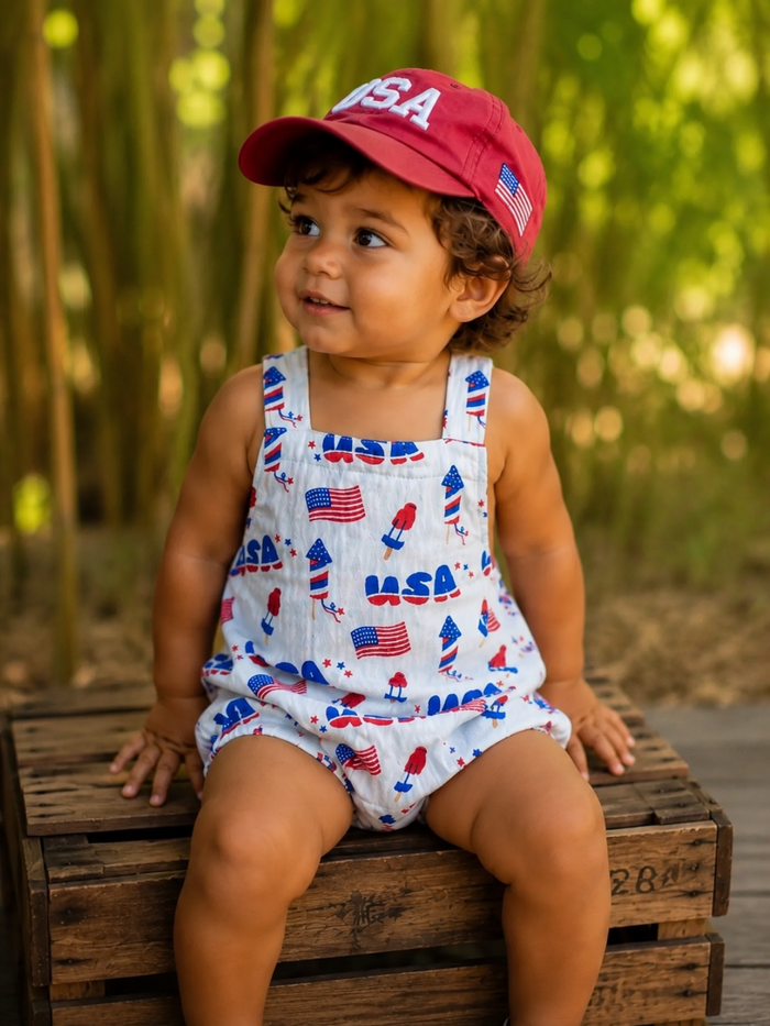 Child in a festive outfit with USA flag prints, sitting on a wooden crate, smiling in a green outdoor setting.