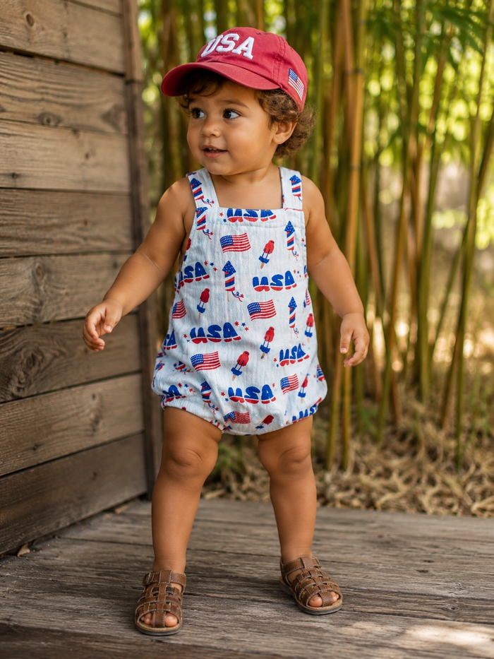 Toddler in an Americana outfit with a red cap, smiling, standing on a wooden surface among greenery.