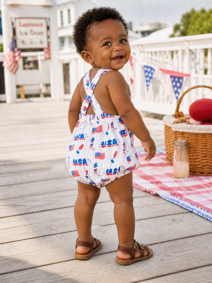 Smiling baby in USA-themed outfit standing on a wooden deck with picnic setup in the background.