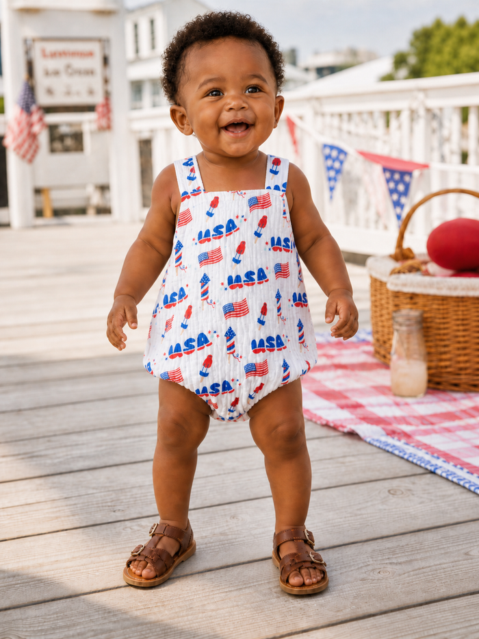 Smiling toddler in a patriotic outfit stands on a wooden deck, surrounded by summer decor and a red picnic blanket.