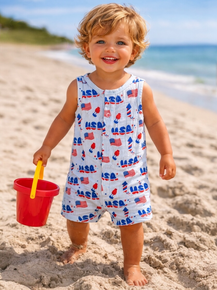 Smiling child in a USA-themed outfit, holding a red bucket while playing on a sandy beach.