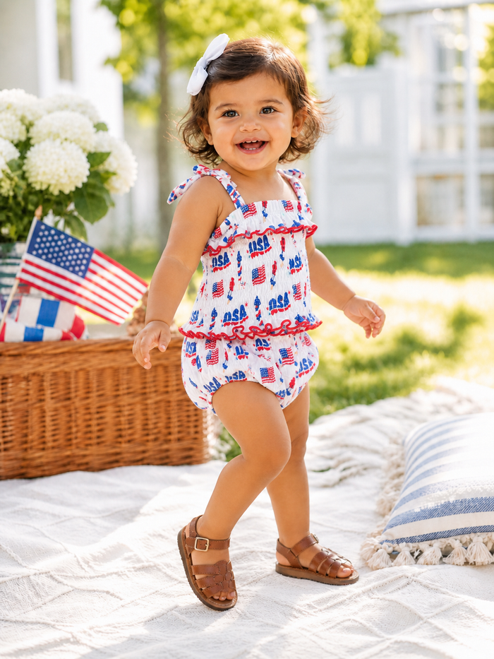 Smiling toddler in patriotic swimwear, enjoying outdoor play with flowers and flags in the background.