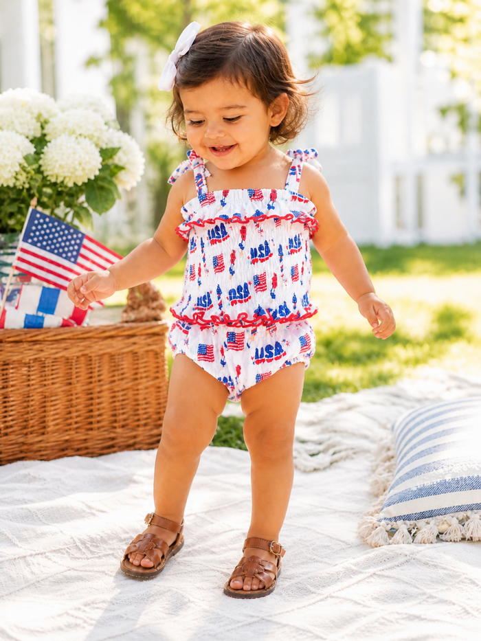 Smiling toddler in a patriotic outfit stands on grass, surrounded by flowers, a basket, and an American flag.