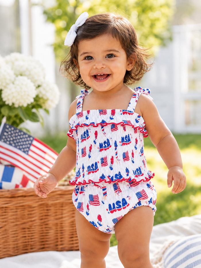 Smiling toddler in patriotic swimsuit with USA print, standing outdoors near flags and flowers.