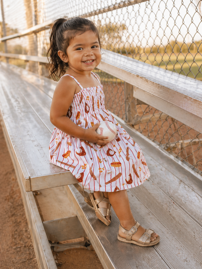 Smiling child in a patterned dress holding a baseball, sitting on metal bleachers at a sunny, outdoor field.