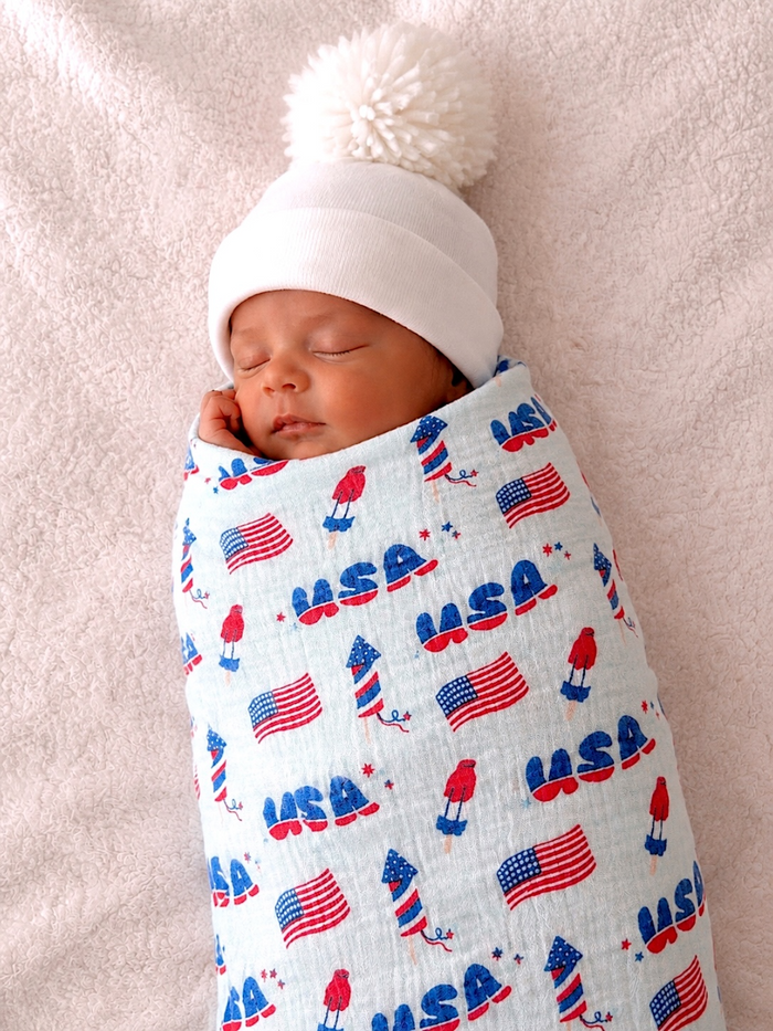 Baby wrapped in a patriotic print swaddle, wearing a white pom-pom hat, peacefully sleeping on a soft background.