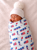 Baby wrapped in a patriotic print swaddle, wearing a white pom-pom hat, peacefully sleeping on a soft background.