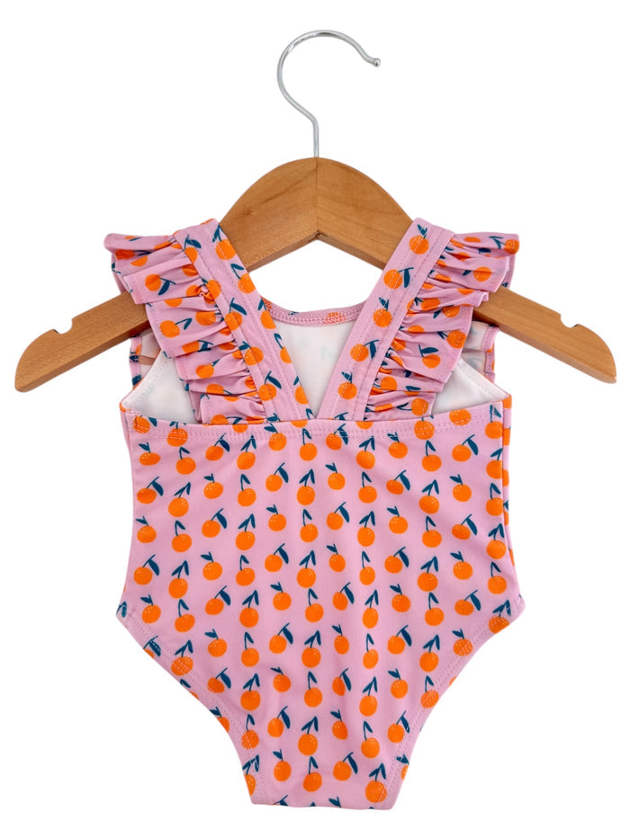 Pink one-piece swimsuit with orange fruit pattern and ruffled straps, hanging on a wooden hanger.