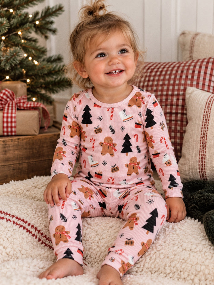 Smiling toddler in festive gingerbread pajamas, sitting on a cozy blanket near a Christmas tree with gifts.