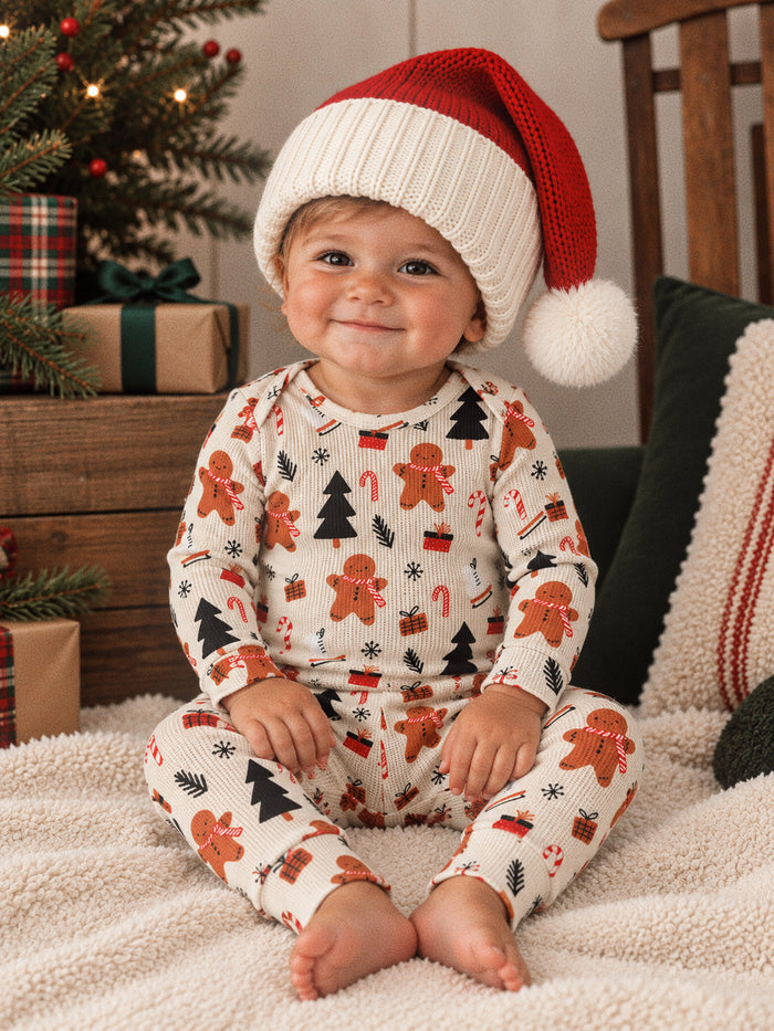 Smiling toddler in festive pajamas with gingerbread, candy canes, and a red Santa hat, sitting by a holiday backdrop.