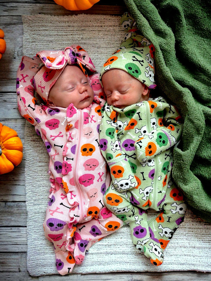 Two newborns dressed in Halloween-themed onesies, sleeping on a textured blanket surrounded by pumpkins.