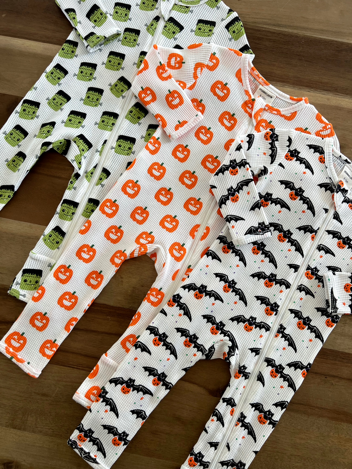 Colorful baby pajamas with Halloween designs: Frankenstein, pumpkins, and bats, arranged on a wooden surface.