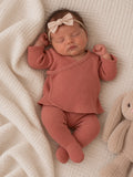 Sleeping baby in a cozy rust outfit with a bow, resting beside a soft stuffed bunny on a textured blanket.