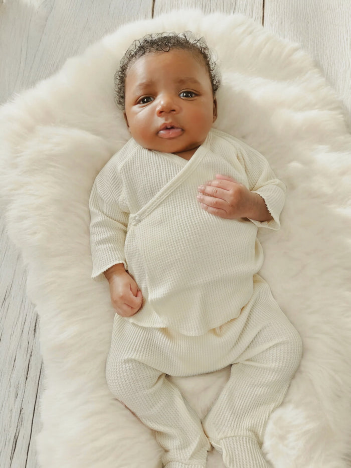 Baby lying on a fluffy white blanket, wearing a cream-colored outfit and looking up with a curious expression.