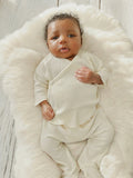 Baby lying on a fluffy white blanket, wearing a cream-colored outfit and looking up with a curious expression.