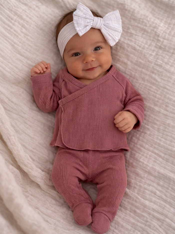 Smiling baby in pink outfit and white headband, lying on soft textured fabric.