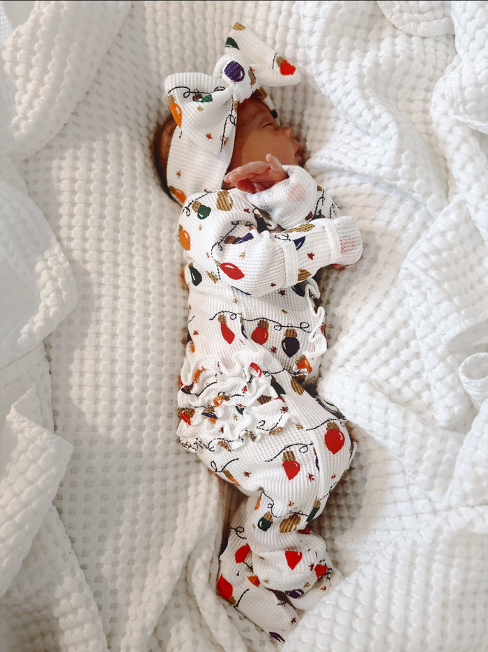 Newborn wearing festive patterned outfit and large bow, peacefully sleeping on white blanket.