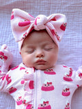 A sleeping baby in a pink outfit with cake patterns and a large bow headband. Soft, peaceful expression on a textured blanket.