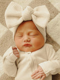 Newborn baby girl asleep in a cozy white outfit with a large bow headband on a textured blanket.